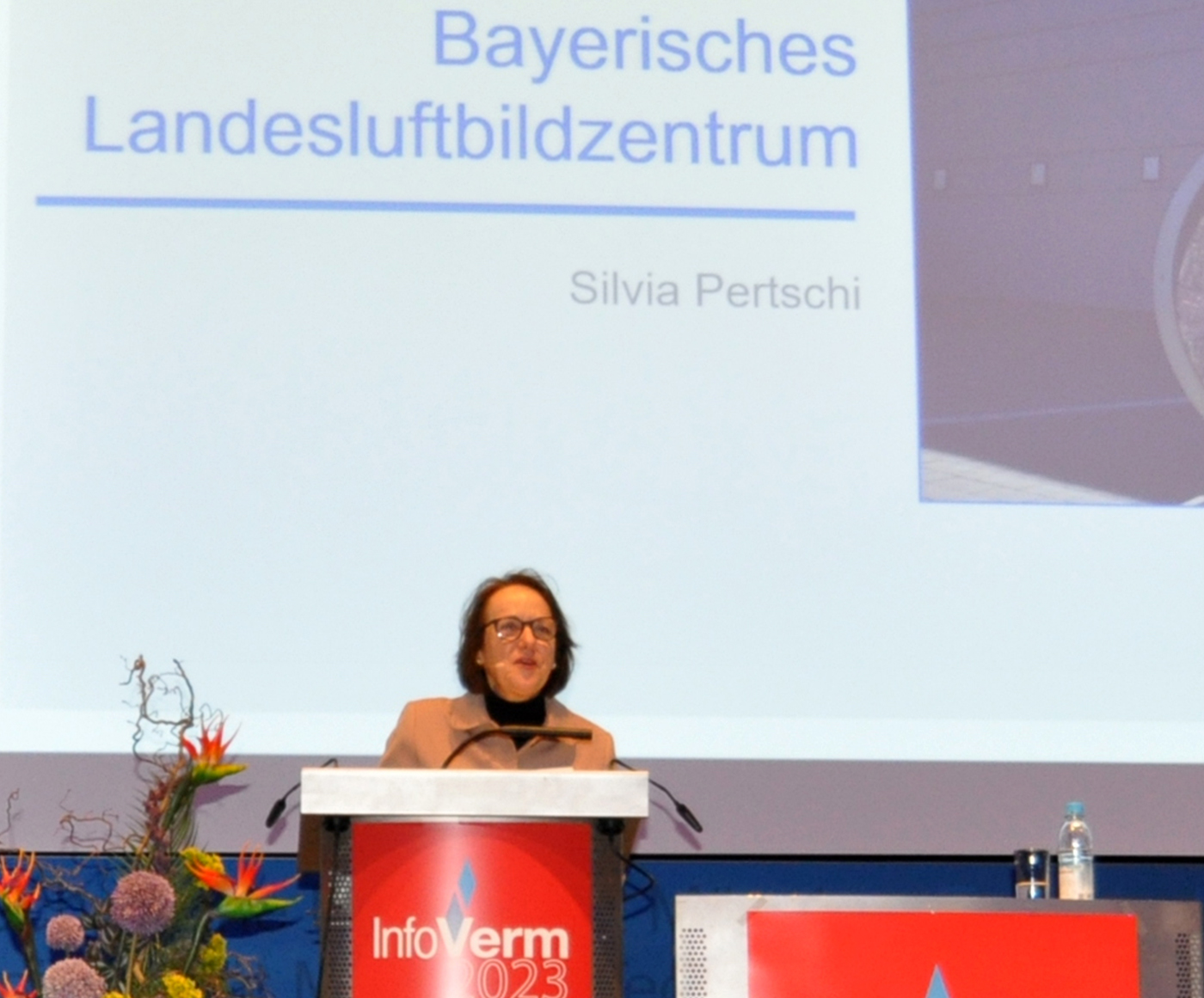 Eine Rednerin steht hinter dem Rednerpult und hält ihren Vortrag. Im Hintergrund steht zu lesen Bayerisches Landesluftbildzentrum.